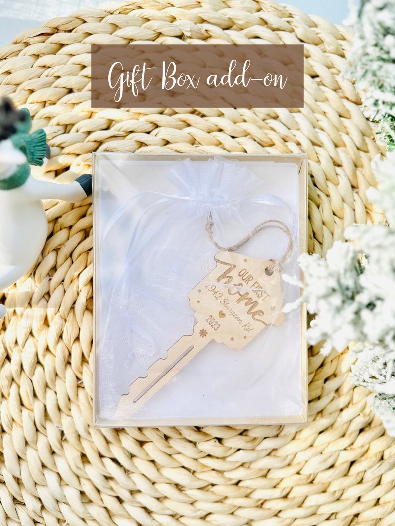 May include: A wooden key-shaped ornament with the text "OUR FIRST HOME" and the address "1942 Sturgeon Rd 2023" inside a clear gift box. The gift box is inside a woven basket. The words "Gift Box add-on" are at the top.