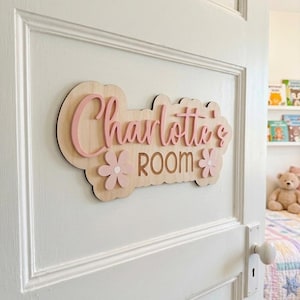 Peut inclure: Une plaque de porte en bois avec une base en bois clair présentant le texte Charlotte's ROOM en lettres roses en relief. La plaque a un bord festonné et est décorée de deux petites fleurs roses de chaque côté du mot ROOM.