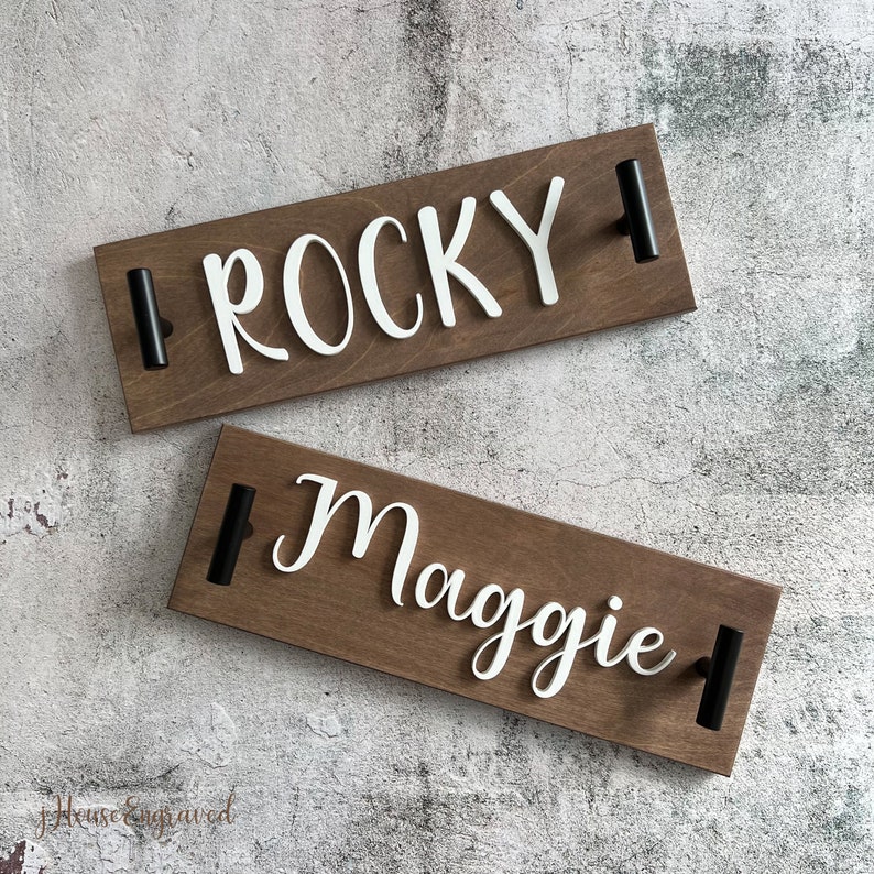 May include: Two wooden signs with the names "Rocky" and "Maggie" in white script. Each sign has a dark brown finish and black handles. The signs are likely for pet identification or home decor.