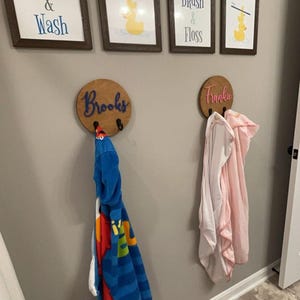 Puede incluir: Dos ganchos de pared circulares de madera con los nombres Brooks y Frankie en letra cursiva. Cada gancho sostiene una toalla con capucha, una azul con estampado de personajes y otra rosa claro. Sobre los ganchos hay cuatro cuadros con patitos.