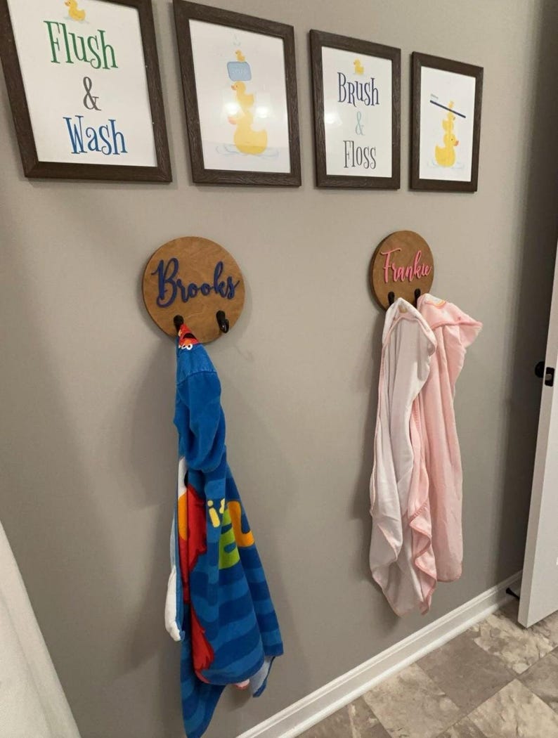 Puede incluir: Dos ganchos de pared circulares de madera con los nombres Brooks y Frankie en letra cursiva. Cada gancho sostiene una toalla con capucha, una azul con estampado de personajes y otra rosa claro. Sobre los ganchos hay cuatro cuadros con patitos.