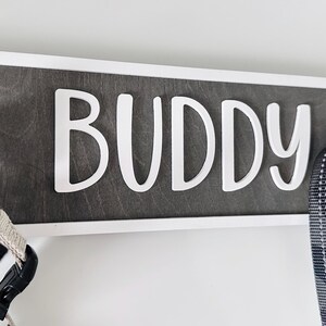 May include: A wooden sign with the name "BUDDY" in white letters. The sign is framed in white and has two black hooks. A black and white dog harness and a leash hang from the hooks.
