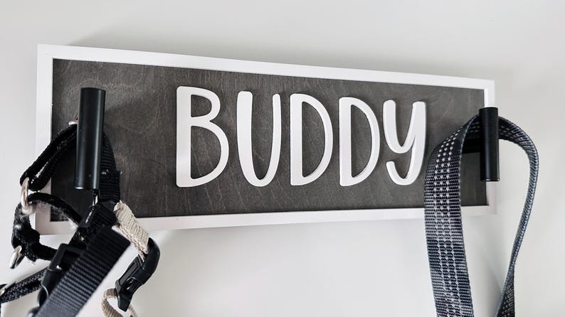May include: A wooden sign with the name "BUDDY" in white letters. The sign is framed in white and has two black hooks. A black and white dog harness and a leash hang from the hooks.