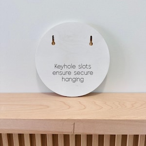 Puede incluir: Una placa de madera clara de forma circular con dos ranuras en forma de cerradura en la parte superior para su montaje. El centro de la placa tiene impreso el texto en negro: Keyhole slots ensure secure hanging. La veta es visible.