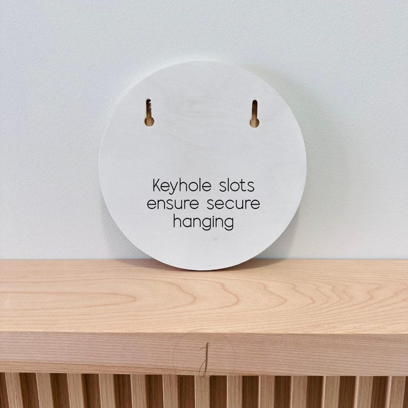 Puede incluir: Una placa de madera clara de forma circular con dos ranuras en forma de cerradura en la parte superior para su montaje. El centro de la placa tiene impreso el texto en negro: Keyhole slots ensure secure hanging. La veta es visible.
