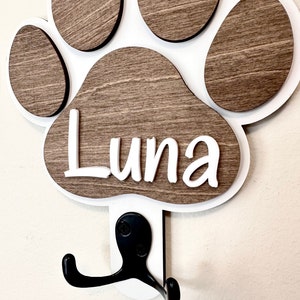 May include: White wooden paw print with a brown wood grain finish. The paw print has the name "Luna" written in white letters. A black metal hook is attached to the bottom of the paw print.