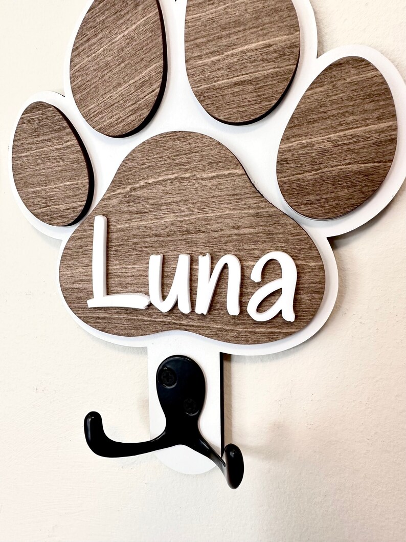 May include: White wooden paw print with a brown wood grain finish. The paw print has the name "Luna" written in white letters. A black metal hook is attached to the bottom of the paw print.