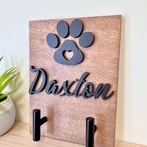 Puede incluir: Porta-correas de perro de madera con dos ganchos negros. El porta-correas presenta una huella de pata negra con un coraz&oacute;n recortado y el nombre "Daxter" en letras negras.