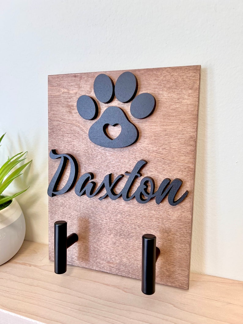 Puede incluir: Porta-correas de perro de madera con dos ganchos negros. El porta-correas presenta una huella de pata negra con un coraz&oacute;n recortado y el nombre "Daxter" en letras negras.