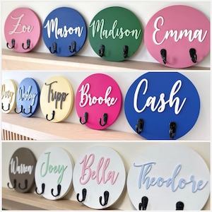 Puede incluir: Una colecci&oacute;n de ganchos de pared circulares de madera con nombres en relieve en estilo cursiva, incluyendo Zoe, Mason, Madison, Emma, Daisy, Wesley, Tripp, Brooke, Cash, Warren, Joey, Bella y Theodore, cada uno con dos ganchos de metal negro.