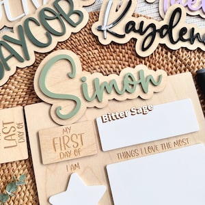 May include: A wooden milestone board featuring the text My First Day Of, I Am, Years Old, Things I Love The Most, and When I Grow Up, I Want To. The board includes a white star shape and blank white rectangular areas for writing with markers.