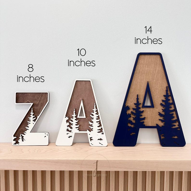 Op de afbeelding: Drie houten letters, Z, A en A, in verschillende maten. De Z is 20 cm hoog, de tweede A is 25 cm en de derde A is 36 cm. Elke letter heeft een uitgesneden ontwerp van een boslandschap, met een natuurlijke houtnerf en witte of marineblauwe verf.