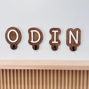 May include: Wooden wall hooks shaped like the letters O, D, I, and N, painted white. The hooks are mounted on a white wall above a wooden shelf.