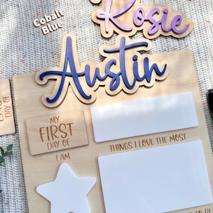 May include: A wooden milestone board featuring the names Rosie and Austin in purple and blue script. The board includes white acrylic sections for writing details like My First Day Of, Things I Love The Most, I Am, and When I Grow Up, I Want To Be.
