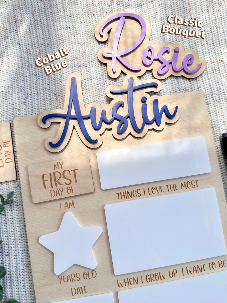 May include: A wooden milestone board featuring the names Rosie and Austin in purple and blue script. The board includes white acrylic sections for writing details like My First Day Of, Things I Love The Most, I Am, and When I Grow Up, I Want To Be.
