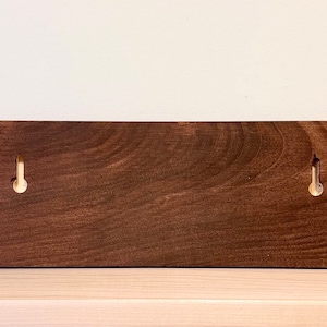May include: A rectangular, stained wooden wall-mounted key holder with two brass hooks and two silver screws. The wood has a rich, dark brown color and a visible wood grain. The hooks are evenly spaced.