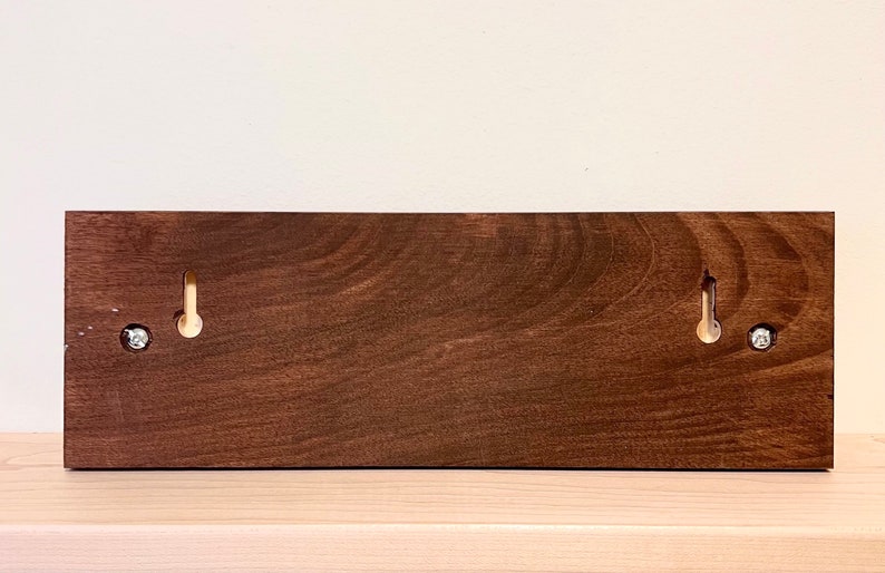 May include: A rectangular, stained wooden wall-mounted key holder with two brass hooks and two silver screws. The wood has a rich, dark brown color and a visible wood grain. The hooks are evenly spaced.
