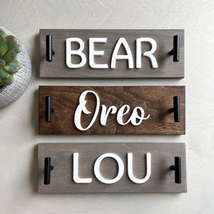 May include: Three wooden name signs with black handles. The top sign is gray with the name "BEAR" in white letters. The middle sign is brown with "Oreo" in white script. The bottom sign is gray with "LOU" in white letters.