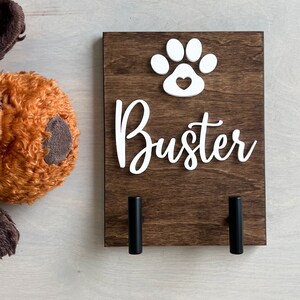 Puede incluir: Un soporte para correa de perro de madera marr&oacute;n con una huella de pata blanca con un coraz&oacute;n en el interior y el nombre "Buster" en letras blancas. Dos ganchos de metal negro est&aacute;n unidos a la parte inferior del soporte.