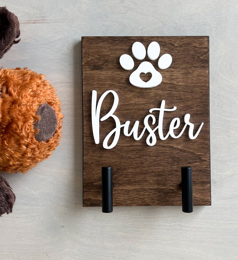 Puede incluir: Un soporte para correa de perro de madera marr&oacute;n con una huella de pata blanca con un coraz&oacute;n en el interior y el nombre "Buster" en letras blancas. Dos ganchos de metal negro est&aacute;n unidos a la parte inferior del soporte.