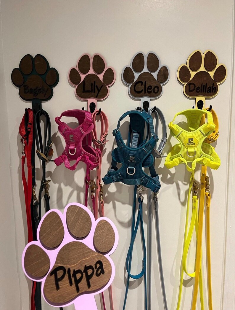 May include: Five wooden paw print hooks with names "Bugsy", "Lily", "Cleo", "Delilah", and "Pippa" are mounted on a white wall. Each hook has a leash and harness hanging from it.