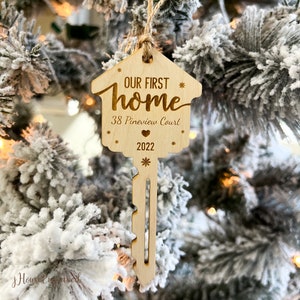May include: A wooden key-shaped Christmas ornament with the text "OUR FIRST home" and "38 Pineview Court 2022". The ornament is hanging on a snowy Christmas tree. The ornament has a house-shaped top and a key-shaped bottom.