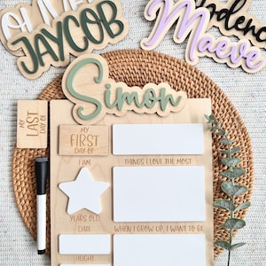 May include: A wooden milestone board featuring the name Simon in green, with sections for My First Day Of, My Last Day Of, I Am, Things I Love The Most, When I Grow Up, I Want To Be, Years Old, Date, and Height, accompanied by a black dry-erase marker.