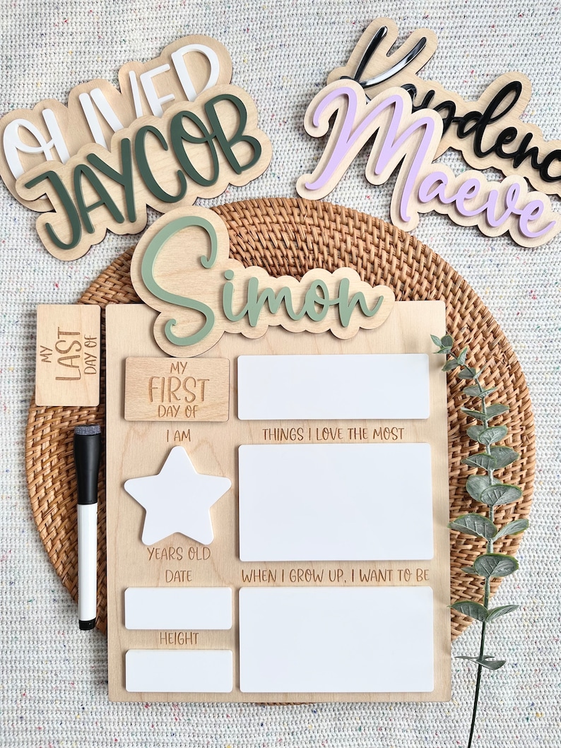 May include: A wooden milestone board featuring the name Simon in green, with sections for My First Day Of, My Last Day Of, I Am, Things I Love The Most, When I Grow Up, I Want To Be, Years Old, Date, and Height, accompanied by a black dry-erase marker.