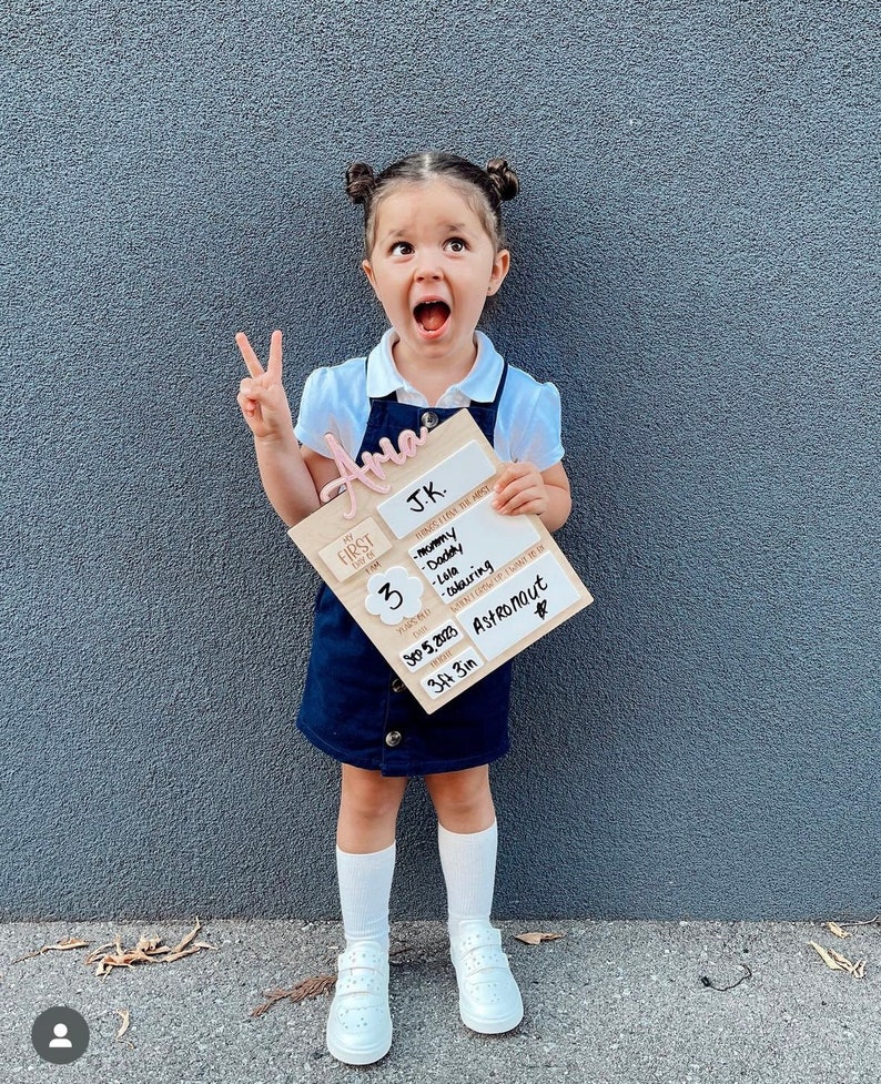 May include: A wooden sign with the words "My Firsts" and "J.K." on it. The sign also includes a list of firsts, such as "Mommy", "Daddy", "Lala", and "Colouring". The sign also includes the date "3/5/2023" and the height "3ft 3in". The sign is held by a young girl with a pink name tag that says "Aria" on it.