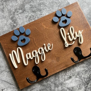 May include: A rectangular wooden wall hook board featuring two blue paw prints with heart-shaped cutouts and the names Maggie and Lily in raised white script lettering. Two black metal double hooks are attached to the stained brown wood surface.