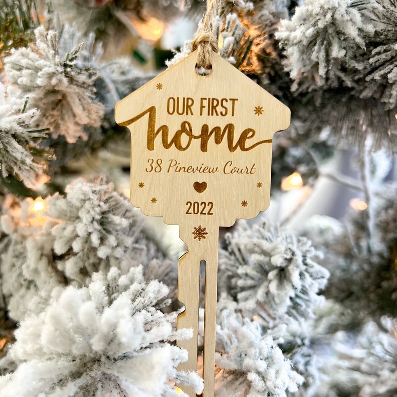 May include: A wooden house-shaped ornament with the text "OUR FIRST home" and "38 Pineview Court 2022". The ornament is key-shaped with a snowflake design at the bottom and is hanging from a natural twine string. The ornament is hanging on a snowy Christmas tree.