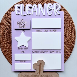 Puede incluir: Un letrero de madera de color lila claro con el nombre "Eleanor" en la parte superior. El letrero tiene espacios para rellenar informaci&oacute;n sobre el primer d&iacute;a de un ni&ntilde;o, incluyendo su edad, altura y lo que quiere ser cuando crezca. El letrero tambi&eacute;n tiene un espacio para una foto y una forma de estrella.