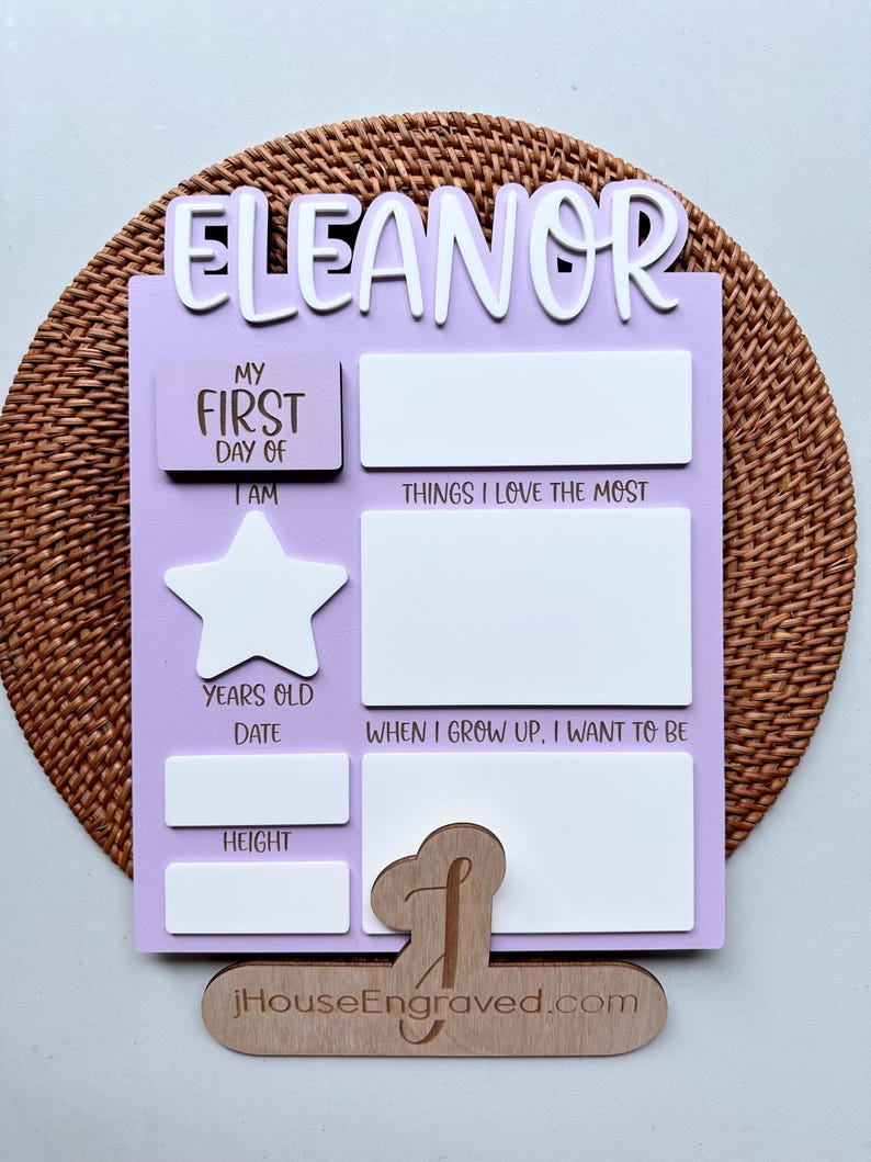 Puede incluir: Un letrero de madera de color lila claro con el nombre "Eleanor" en la parte superior. El letrero tiene espacios para rellenar informaci&oacute;n sobre el primer d&iacute;a de un ni&ntilde;o, incluyendo su edad, altura y lo que quiere ser cuando crezca. El letrero tambi&eacute;n tiene un espacio para una foto y una forma de estrella.