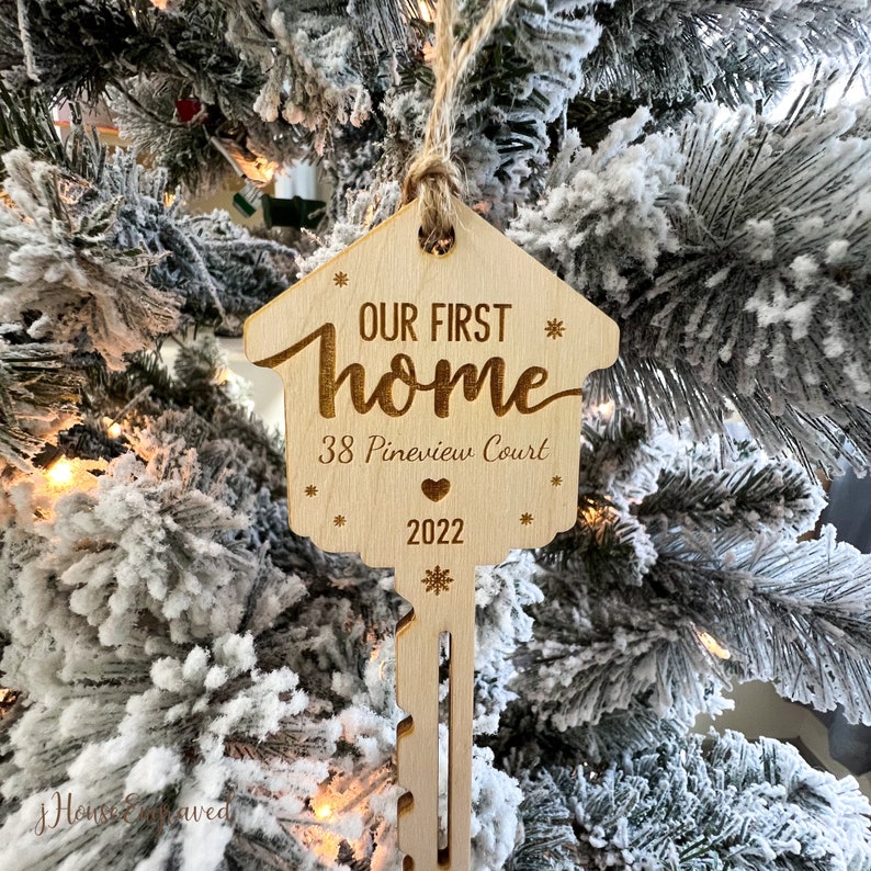 May include: A wooden house-shaped ornament with the text "OUR FIRST HOME", address, and the year 2022. The ornament is hanging from a twine string. The background is a snow-covered Christmas tree.