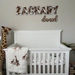 Op de afbeelding: Een witte wieg met een beddengoedset met giraffenthema en een pluche giraf. Boven de wieg staat de naam "ZACHARY" in houten letters met een safari-dierenontwerp en de naam "daniel" eronder.
