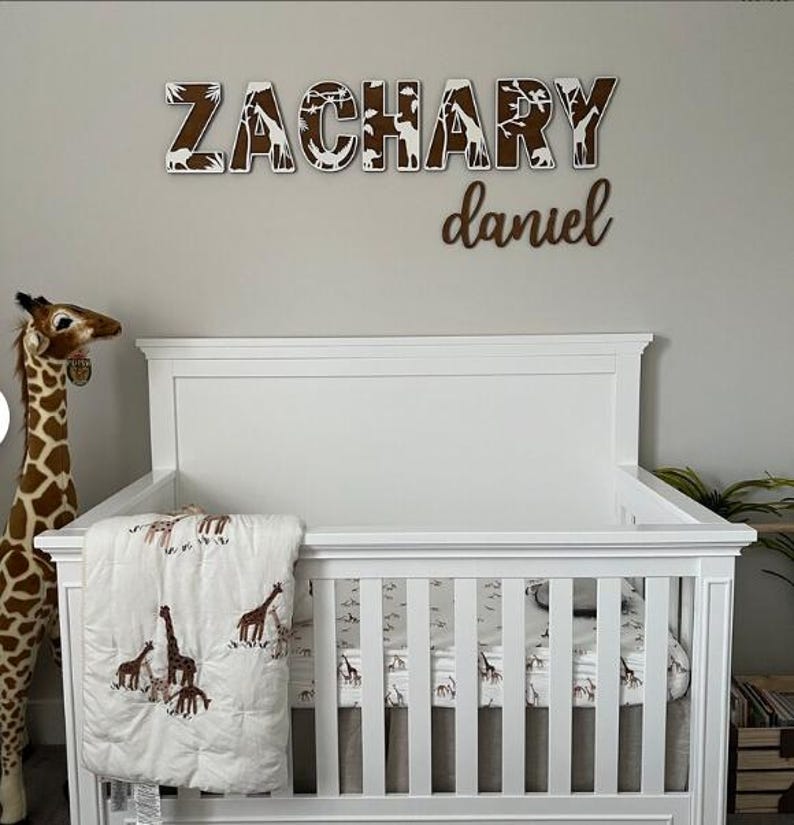 Op de afbeelding: Een witte wieg met een beddengoedset met giraffenthema en een pluche giraf. Boven de wieg staat de naam "ZACHARY" in houten letters met een safari-dierenontwerp en de naam "daniel" eronder.