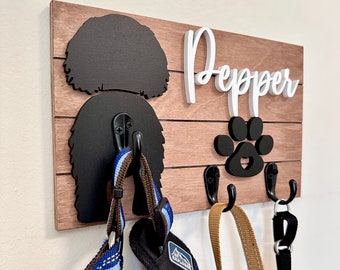Peut inclure: Un porte-laisses et colliers pour chien en bois avec une silhouette noire de tête de chien et une empreinte de patte. Le porte-laisses a trois crochets et le nom "Pepper" est écrit en lettres blanches sur le bois.