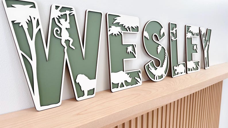 Op de afbeelding: Houten naambord met de naam "WESLEY" in wit en groen. Elke letter heeft een silhouet van een jungle-dier of plant. Het bord staat op een lichtgekleurde houten plank.