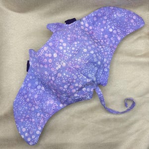 Puede incluir: Un sombrero para el sol de tela con forma de manta raya, con un estampado de lunares blancos y morados sobre un fondo azul. El sombrero tiene una visera curva y una correa con hebilla.