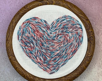 May include: A heart-shaped embroidery piece with a swirling pattern of red, white, and blue threads. The artwork is set in a round, wooden embroidery hoop with a dark brown finish. The heart is centered on a white fabric background.