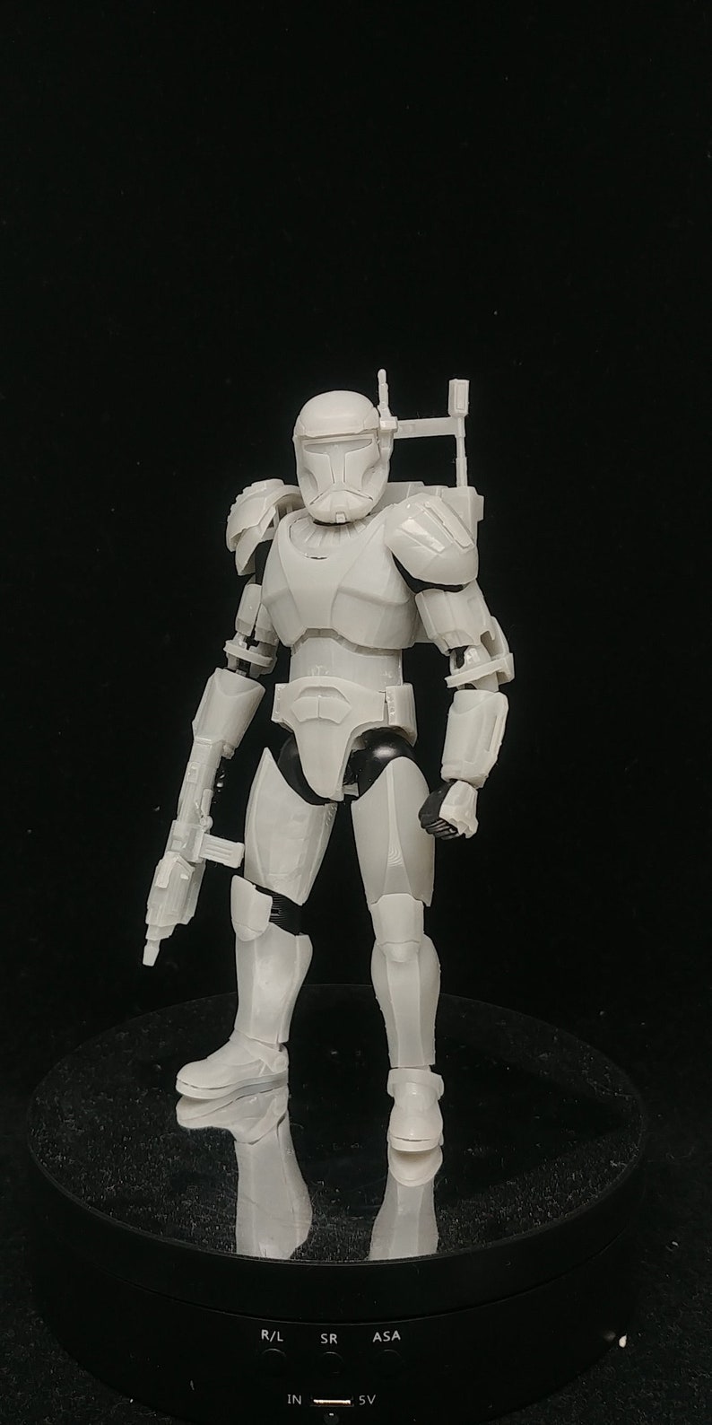 May include: A light gray action figure of a soldier in armor, holding a blaster rifle. The figure has a helmet, shoulder pads, and a backpack. The figure is standing on a black rotating display stand.