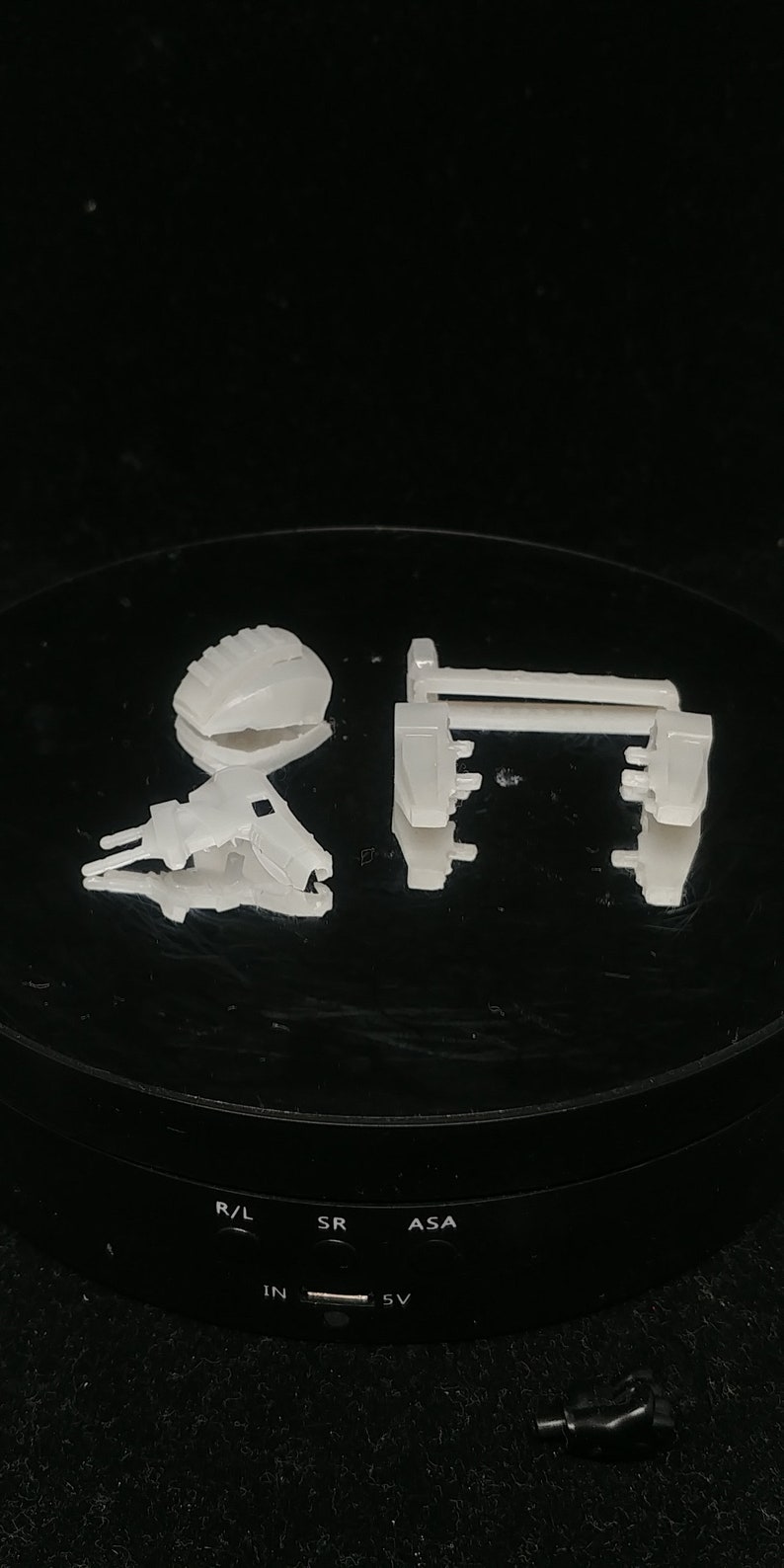 May include: A collection of small, off-white plastic components, possibly for a larger device, are arranged on a black surface. The pieces include a rectangular frame, a curved piece, and a complex, angled structure.