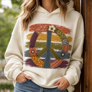 May include: A cream-colored hooded sweatshirt featuring a large peace sign graphic on the front. The peace sign is composed of various patterned fabric-like segments in shades of blue, green, orange, and yellow, accented with floral details.