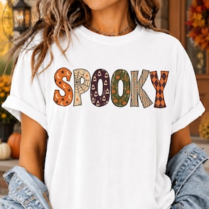 May include: A white short-sleeved t-shirt featuring the word SPOOKY printed across the chest. Each letter has a unique pattern including orange stars and moons, a spider web, candy corn, green pumpkins, black stripes, and orange diamonds.
