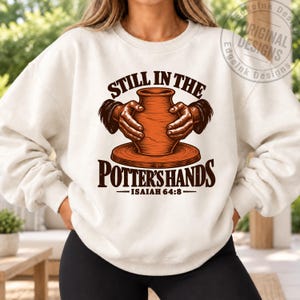 May include: A cream-colored crewneck sweatshirt featuring a graphic of two hands shaping a clay pot on a pottery wheel. The text reads "STILL IN THE POTTER'S HANDS" and "ISAIAH 64:8" in dark brown lettering. The design is centered on the chest.