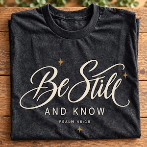 Op de afbeelding: Een antracietgrijs T-shirt met ronde hals en de crèmekleurige tekst "Be Still AND KNOW" in een combinatie van script- en schreefloze lettertypes. Het ontwerp bevat drie kleine gouden vierpuntige sterren en de tekst "PSALM 46:10" eronder.