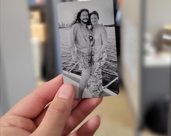 May include: A black and white photo of a couple on a boat. The man has long hair and is wearing a button-down shirt and shorts. The woman is wearing a tank top and a skirt. They are both smiling.