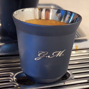 May include: A dark blue ceramic cup with a silver rim and a silver inscription that reads "G.M.". The cup is filled with a light brown liquid.
