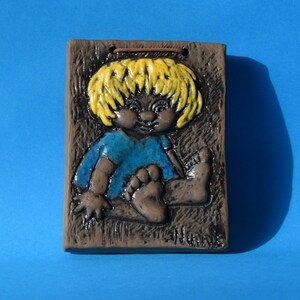 Può includere: Una targa da parete in ceramica con un bambino con i capelli gialli, che indossa una camicia blu e siede con le gambe incrociate. La targa ha uno sfondo marrone con una texture a grana di legno.
