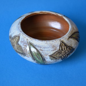 Op de afbeelding: Een handgemaakte keramische kom met een lichtbeige buitenkant en een warmbruine binnenkant. De kom heeft reliëf bloemen- en bladerontwerpen in groen- en bruintinten. De kom heeft een diameter van ongeveer 15 cm.